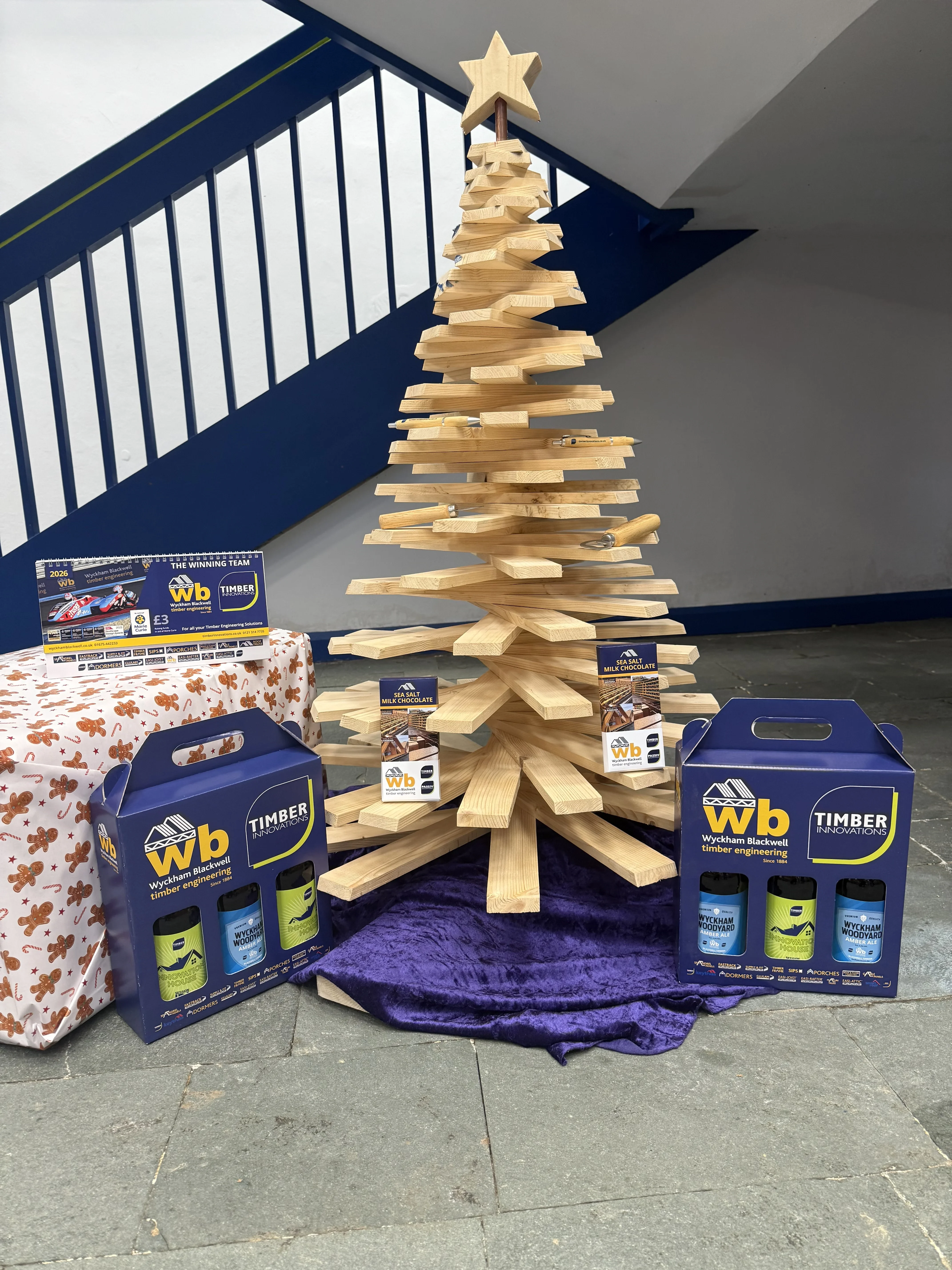 A Christmas tree made out of wood with WB and Timber Innovation beer packages below it.