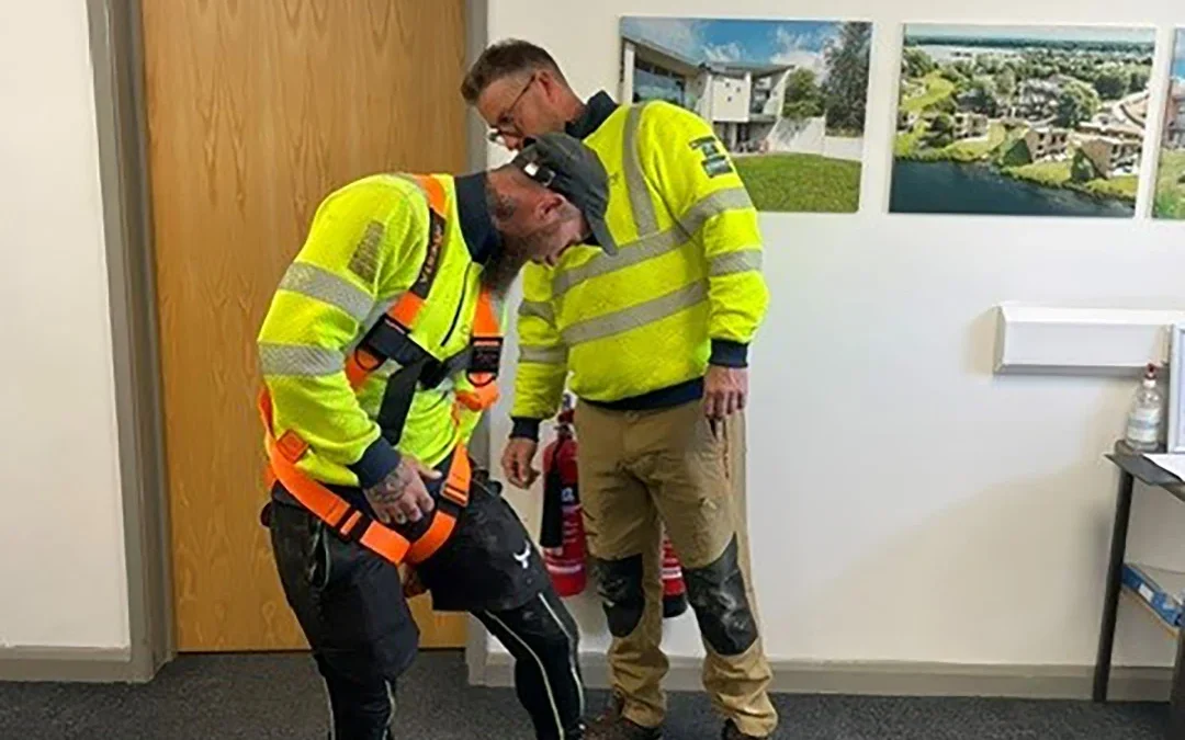 Two construction workers in hi-vis jackets in an office, one adjusting a safety harness on the other; Glosford SIPS logo.