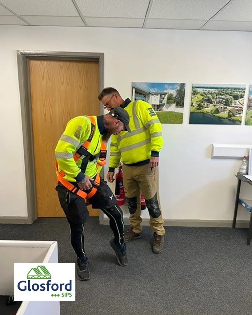 Two construction workers in hi-vis jackets in an office, one adjusting a safety harness on the other; Glosford SIPS logo.