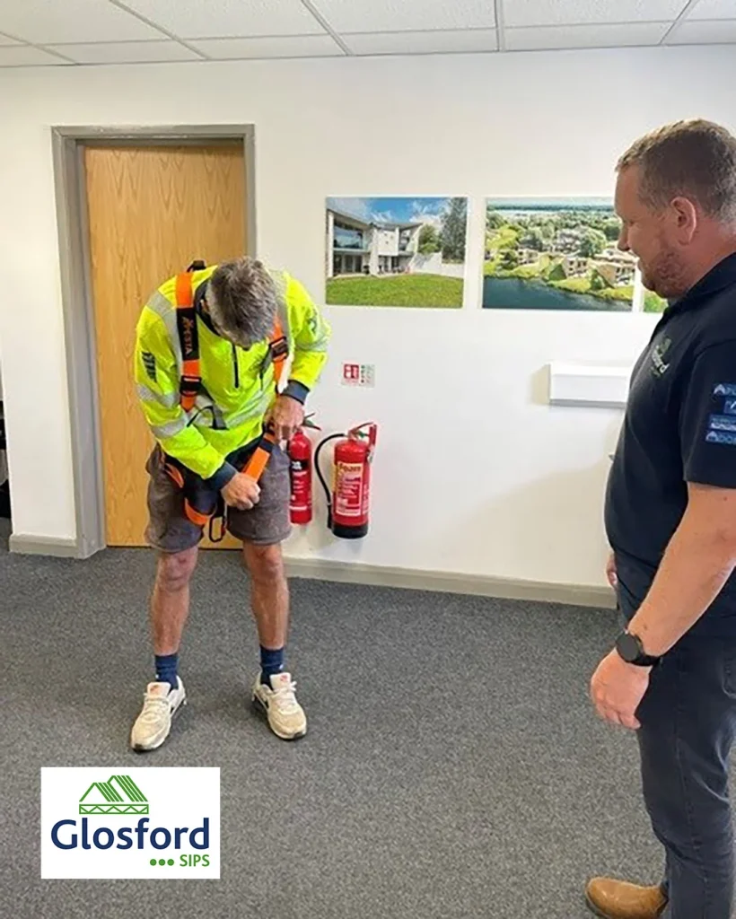 Worker in high-visibility jacket adjusts safety harness as colleague watches, fire extinguishers on wall and Glosford SIPS logo.