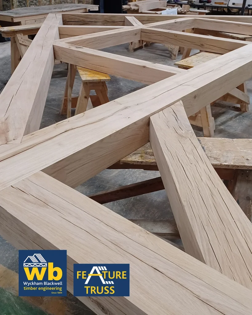 Large oak timber feature truss with intersecting beams resting on sawhorses in a workshop, Wyckham Blackwell logo in bottom right.