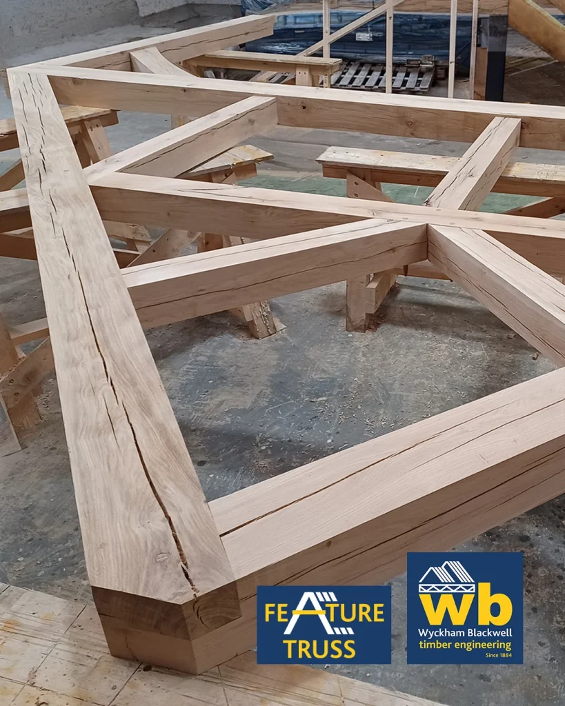 Large oak timber feature truss with intersecting beams resting on sawhorses in a workshop, Wyckham Blackwell logo in bottom right.