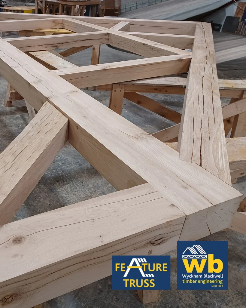 Large oak timber feature truss with intersecting beams resting on sawhorses in a workshop, Wyckham Blackwell logo in bottom right.