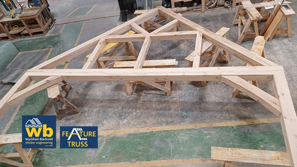 Large oak timber feature truss with intersecting beams resting on sawhorses in a workshop, Wyckham Blackwell logo in bottom right.