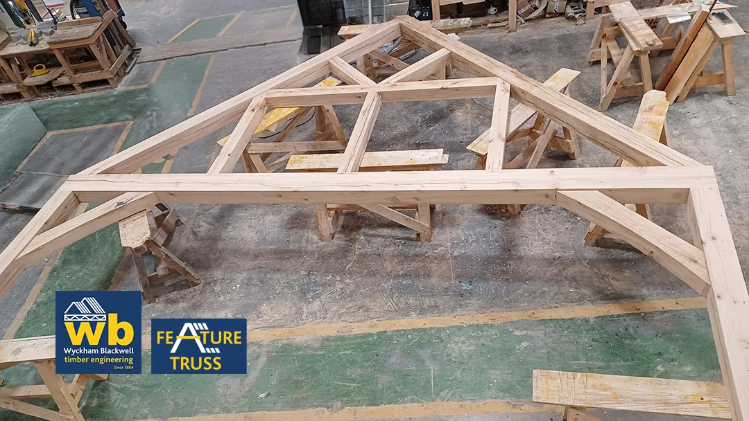Large oak timber feature truss with intersecting beams resting on sawhorses in a workshop, Wyckham Blackwell logo in bottom right.