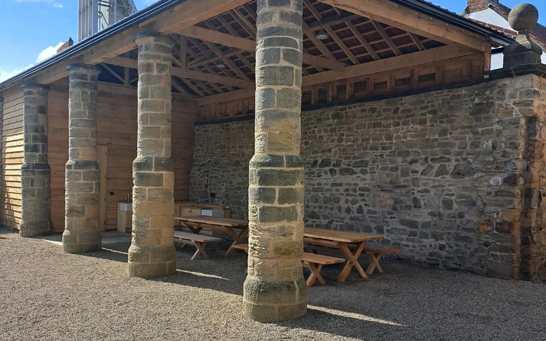 Open timber pavilion with exposed timber trusses, stone columns and wall, picnic benches and service counters; Crendon logo in bottom right