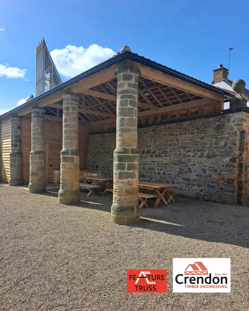 Open timber pavilion with exposed timber trusses, stone columns and wall, picnic benches and service counters; Crendon logo in bottom right