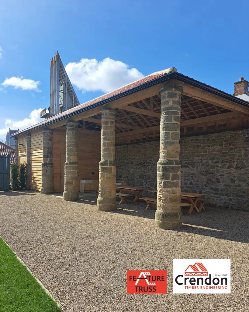 Open timber pavilion with exposed timber trusses, stone columns and wall, picnic benches and service counters; Crendon logo in bottom right