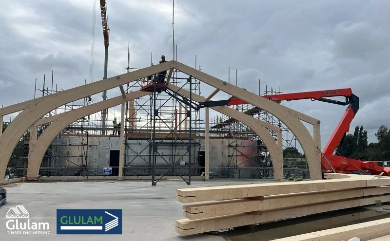 Glulam timber arches being installed at a construction site, workers on lifts and stacked beams under a cloudy sky.