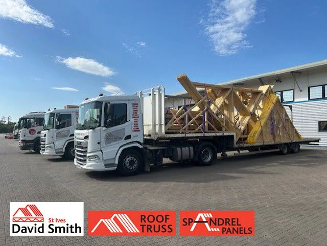 Three white trucks by a warehouse; one flatbed carries wooden roof trusses, branded with David Smith and roof truss logos.