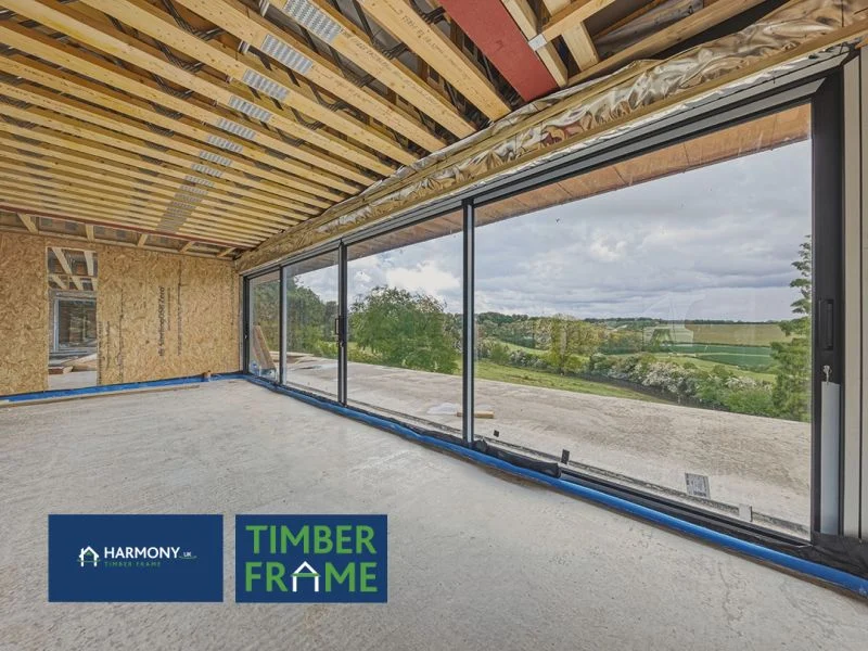 Unfinished timber-frame room with exposed joists and floor-to-ceiling glass overlooking rolling countryside; company logos.
