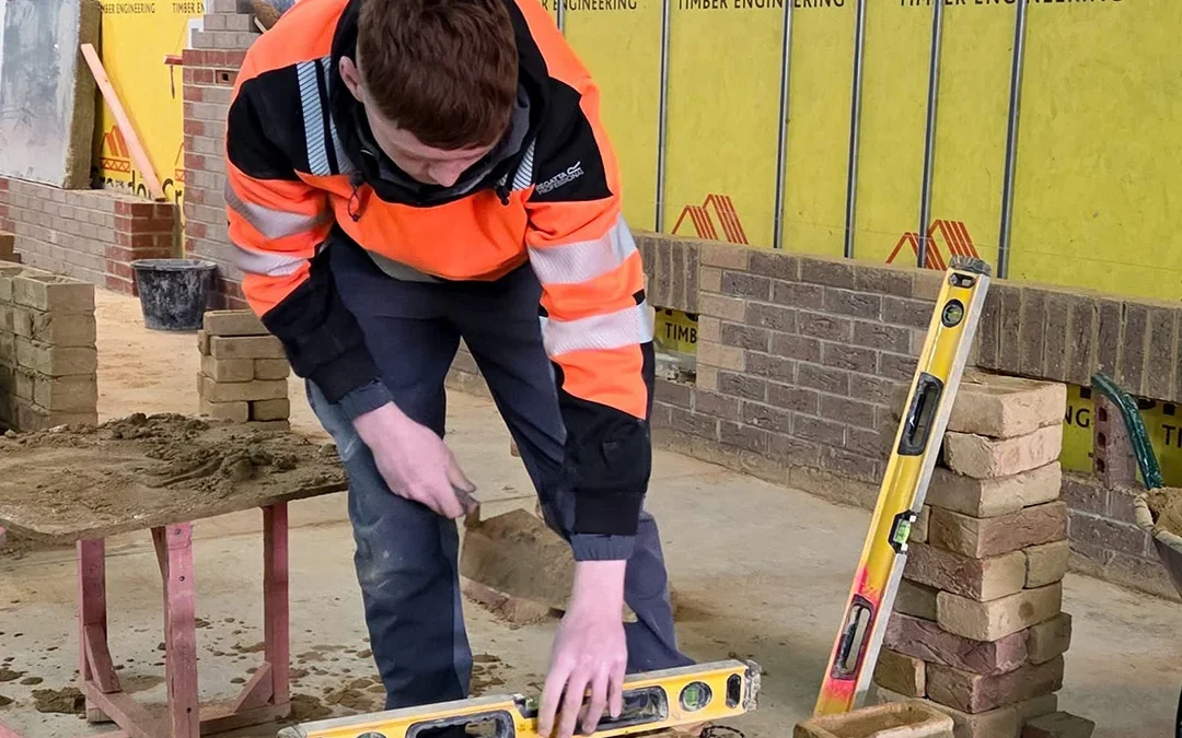 Worker in orange hi-vis jacket laying bricks with trowel and spirit level in indoor masonry workshop with Crendon signage.
