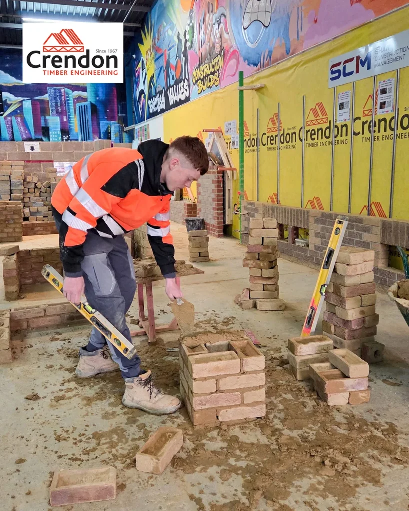 Worker in orange hi-vis jacket laying bricks with trowel and spirit level in indoor masonry workshop with Crendon signage.