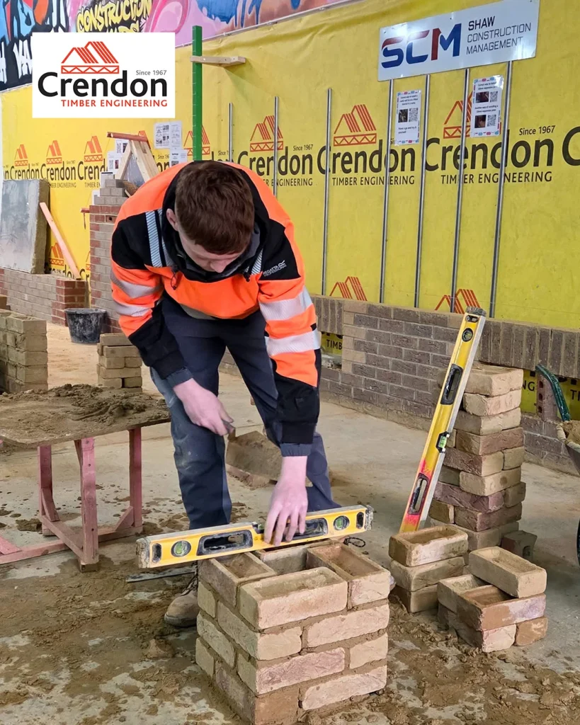 Worker in orange hi-vis jacket laying bricks with trowel and spirit level in indoor masonry workshop with Crendon signage.