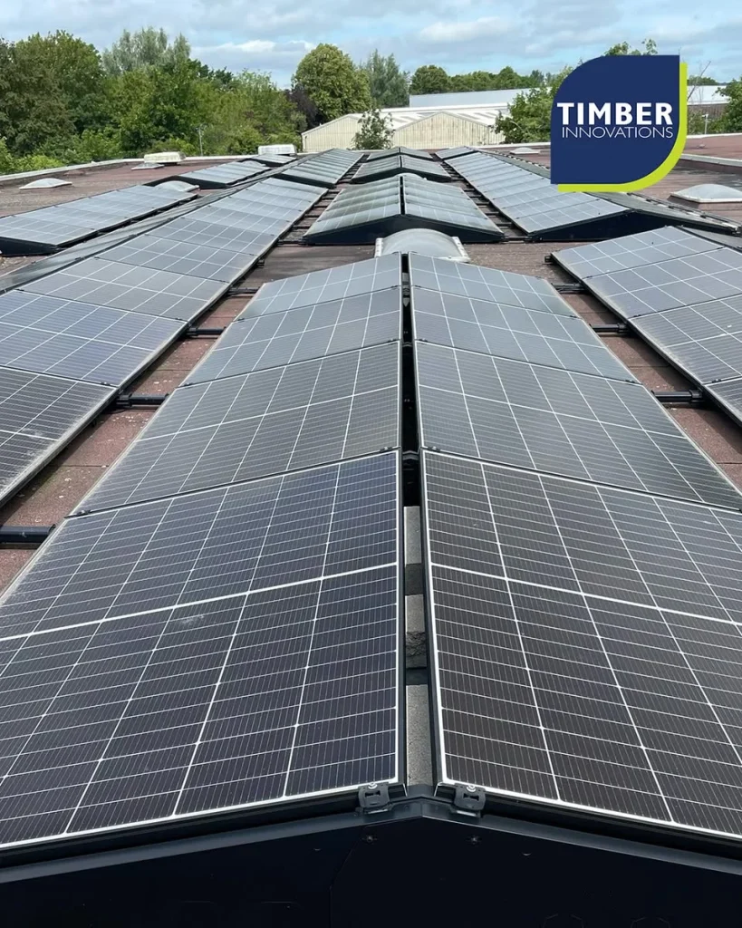Multiple rows of rooftop solar panels on a commercial roof, trees and buildings in the background, Timber Innovations logo.