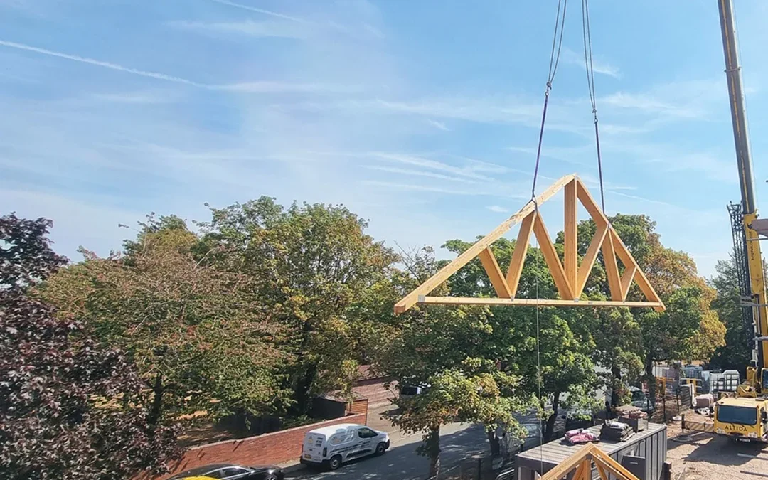 Crane lifts a wooden Roof Truss above a flatbed lorry at a construction site, Wyckham Blackwell logos at top.